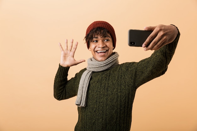 Retrato de un joven africano feliz vestido con ropa de otoño aislado sobre pared beige, tomando un selfie, saludando