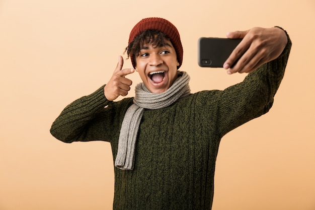 Retrato de un joven africano feliz vestido con ropa de otoño aislado sobre pared beige, tomando un selfie