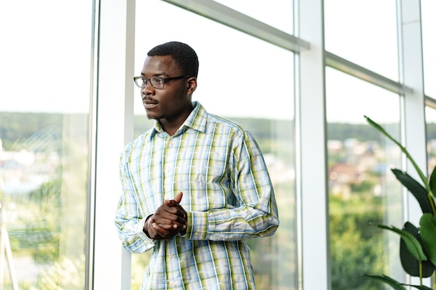 Retrato de un joven africano pensativo con gafas de pie cerca de la ventana