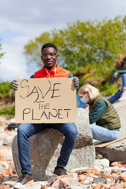 Retrato de joven africano sosteniendo cartel y mirando a cámara mientras está sentado en la piedra al aire libre