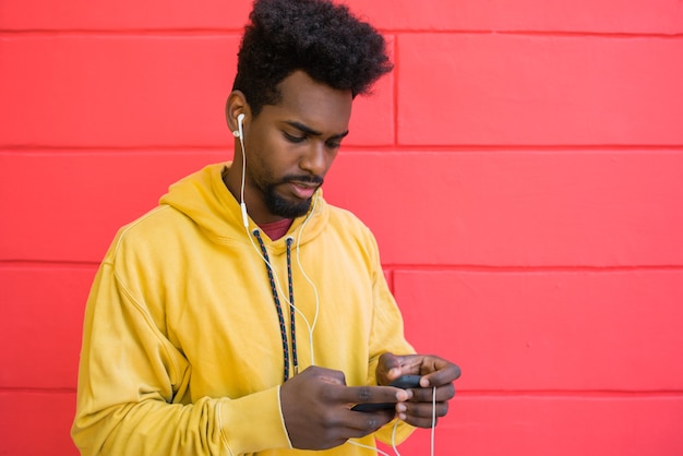 Retrato de joven afro usando su teléfono móvil con auriculares contra la pared roja. Concepto de tecnología y estilo de vida.