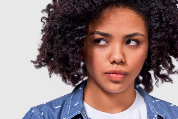 El retrato de una joven afroamericana loca tiene una expresión facial triste y viste ropa casual aislada sobre un fondo blanco de estudio Mujer molesta mirando a un lado sobre una pared gris