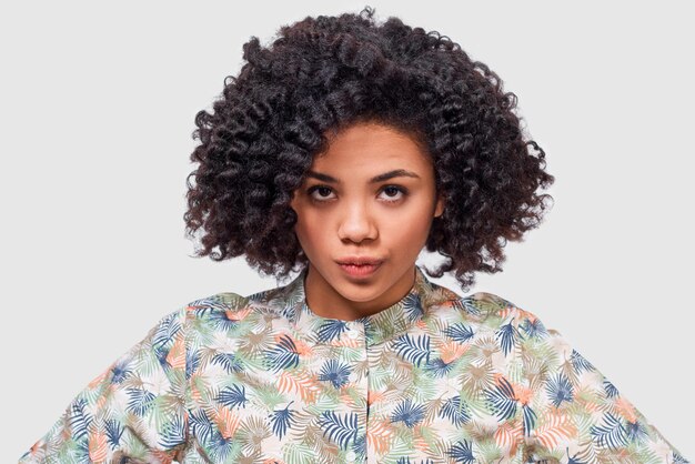 Retrato de una joven afroamericana seria que usa una camisa floral mirando a la cámara tiene poses de expresión frustrada en el estudio La mujer afroamericana tiene una expresión enojada
