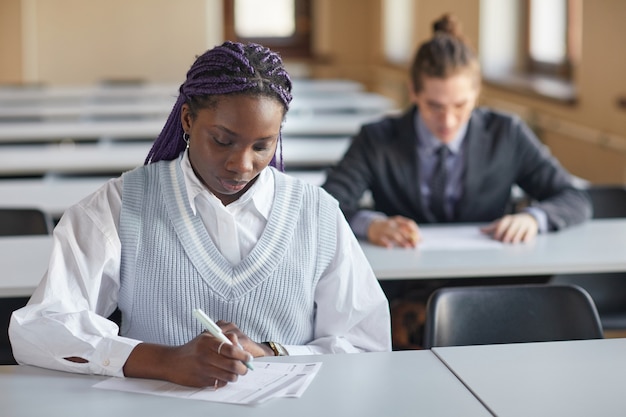 Retrato de joven afroamericana vistiendo uniforme escolar mientras toma el examen en el auditorio de la universidad, espacio de copia