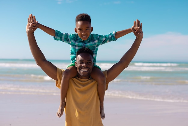 Retrato de un joven afroamericano alegre con los brazos levantados llevando a su hijo sobre los hombros en la playa