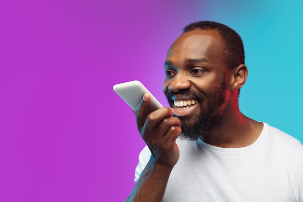 Retrato de joven afroamericano sobre fondo de estudio degradado en neón. Hermoso modelo masculino de estilo casual, camisa blanca. Concepto de emociones humanas, expresión facial, ventas, publicidad.