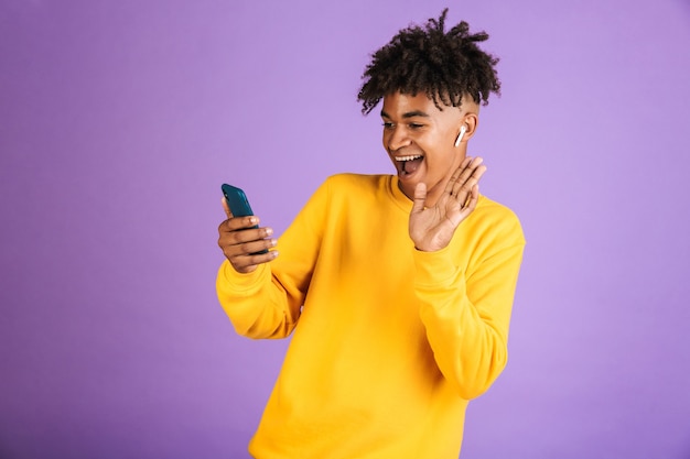 Retrato de joven afroamericano sonriendo y agitando la mano en el teléfono inteligente, usando auriculares bluetooth, aislado sobre fondo violeta