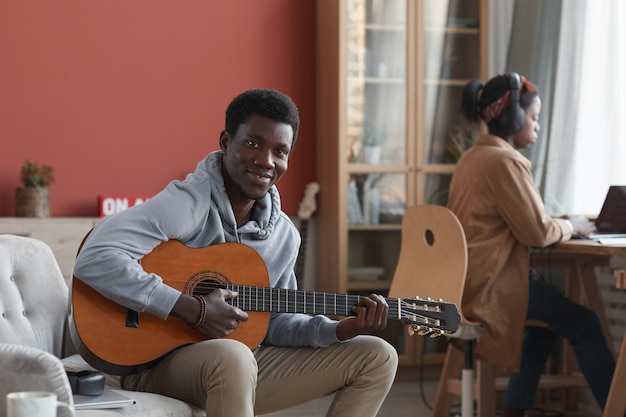 Retrato de joven afroamericano tocando la guitarra acústica y sonriendo a la cámara mientras está sentado en el sofá en el estudio de grabación, espacio de copia