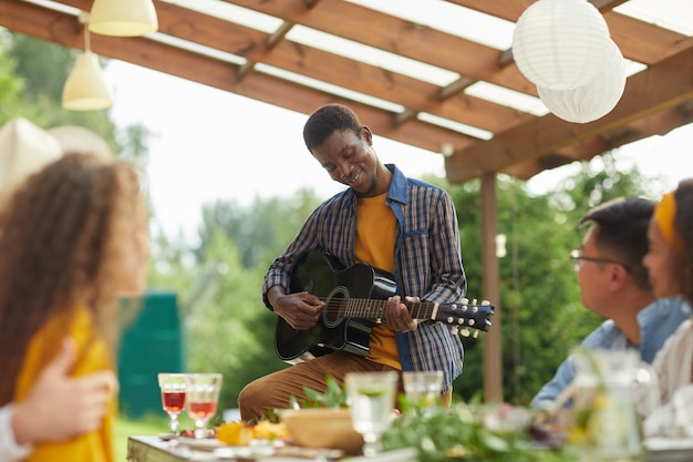 Retrato de joven afroamericano tocando la guitarra mientras está de pie junto a la mesa y disfruta de una cena con amigos al aire libre en la fiesta de verano