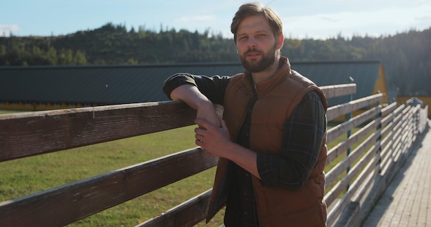 Retrato de un joven agricultor apoyado en una valla contra el fondo de los edificios de la granja en un día soleado