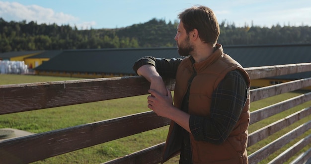 Retrato de un joven agricultor apoyado en una valla contra el fondo de los edificios de la granja en un día soleado