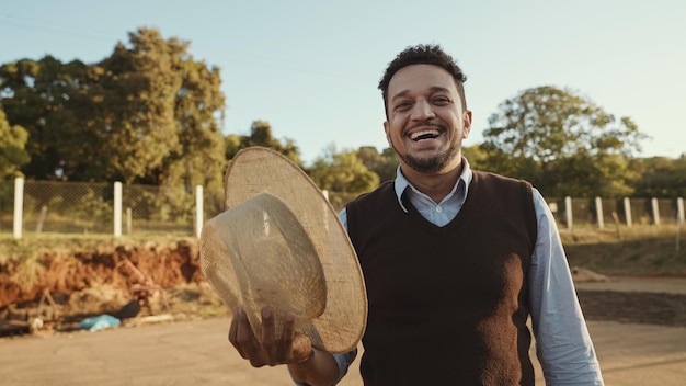 Retrato de un joven agricultor latino con un pantalones informal quitándose el sombrero para la cámara en el fondo de la granja