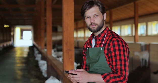 Retrato de un joven agricultor sentado en un establo de cabras y enviando mensajes de texto en un teléfono inteligente