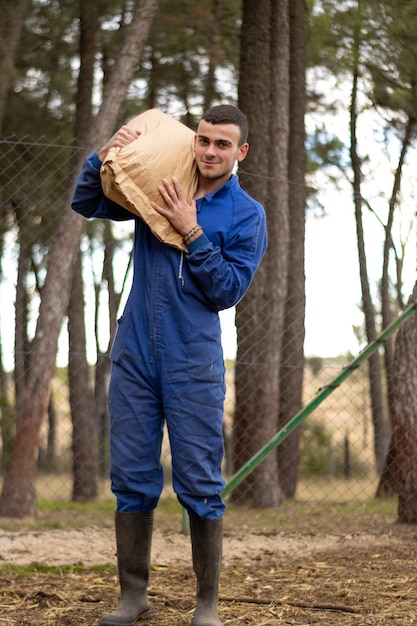 Retrato del joven agricultor sonriente con un saco de alimento en su hombro en su granja y mirando a la cámara., Fondo de naturaleza vertical