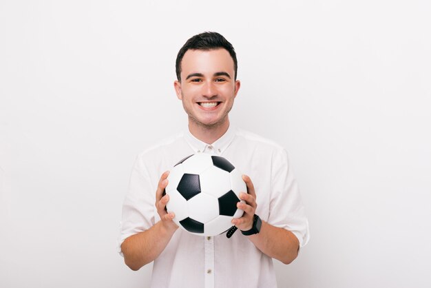 Retrato de joven alegre con balón de fútbol y mirando a la cámara, de pie sobre la pared blanca