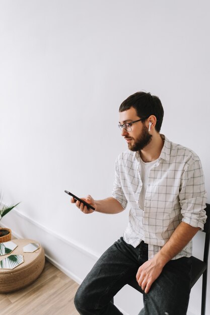 Retrato de joven alegre caucásico en gafas y auriculares mediante teléfono móvil.