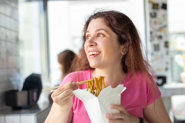 Retrato de una joven alegre comiendo fideos chinos en un café y mirando por la ventana. El concepto de cocina asiática saludable.