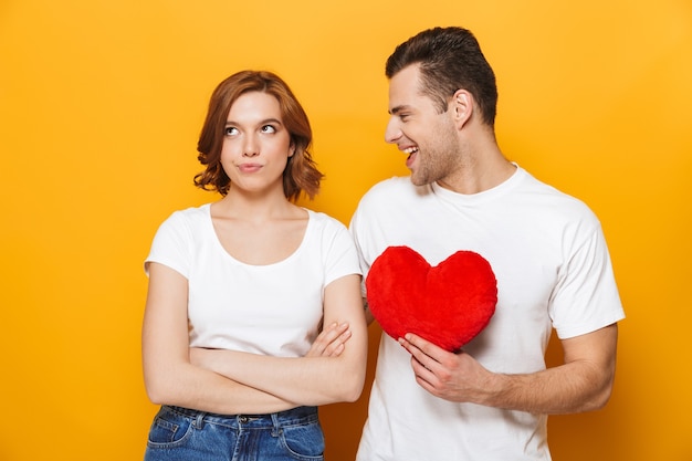 Retrato de un joven alegre dando corazón rojo a la novia decepcionada que se encuentran aisladas sobre la pared amarilla