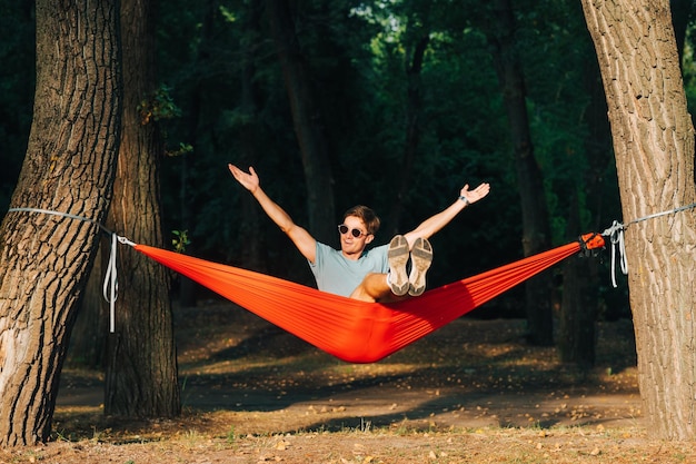Retrato de un joven alegre con gafas de sol sentado en una hamaca en el parque