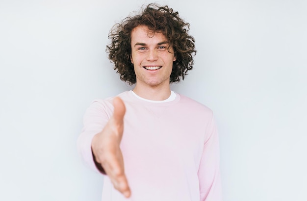 Retrato de un joven alegre con un moderno peinado rizado sonriendo ampliamente mientras estira la mano a la cámara con un gesto de apretón de manos sobre fondo blanco Concepto de personas y lenguaje corporal