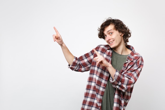 Retrato de joven alegre en ropa casual mirando a cámara apuntando con el dedo índice a un lado aislado sobre fondo de pared blanca en estudio. Concepto de estilo de vida de emociones sinceras de personas. Simulacros de espacio de copia.