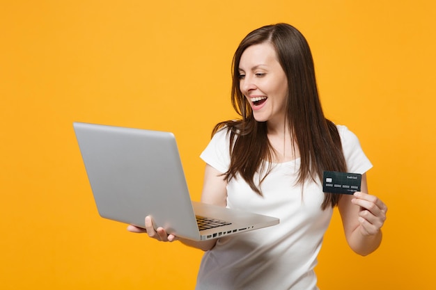 Retrato de una joven alegre con ropa informal blanca sosteniendo un ordenador portátil, tarjeta bancaria de crédito aislada en un fondo de pared naranja amarillo en el estudio. Concepto de estilo de vida de las personas. Simulacros de espacio de copia.