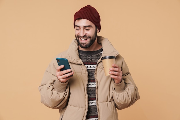 Retrato de joven alegre en ropa de invierno con taza de café y teléfono móvil aislado en beige