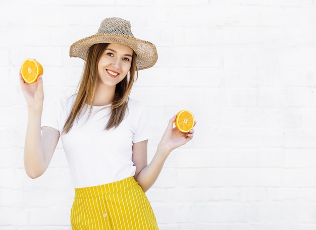 Retrato de una joven alegre con sombrero sosteniendo dos rodajas de naranja en la pared blanca