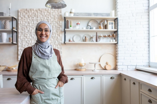 Retrato de una joven ama de casa árabe con hiyab parada en la cocina cerca de la mesa con delantal sosteniendo