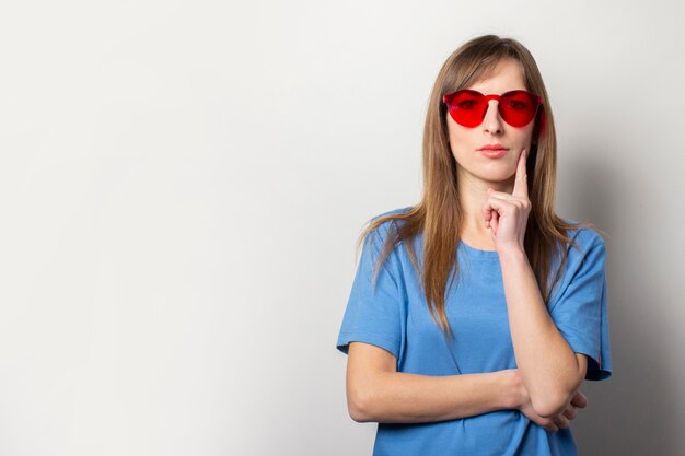 Retrato de una joven amigable en una camiseta azul casual, gafas rojas, sostiene su mano en la barbilla en la luz. Cara emocional Gesto para pensar, planificar