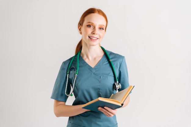 Retrato de una joven y amigable doctora en uniforme verde con estetoscopio sosteniendo un libro médico de pie y mirando la cámara de pie sobre fondo blanco aislado en el estudio