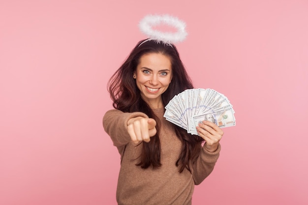 Retrato de una joven angelical feliz con halo sobre la cabeza sosteniendo billetes en dólares y señalando a la cámara eligiendo al ganador de la lotería alentando a ganar mucho dinero en un estudio interior con fondo rosa