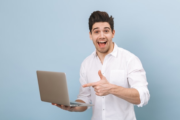 Retrato de un joven apuesto alegre que se encuentran aisladas en azul, trabajando en equipo portátil