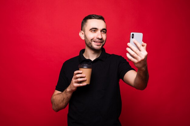 Retrato de un joven apuesto alegre que se encuentran aisladas sobre fondo rojo, sosteniendo una taza de café para llevar, tomando un selfie
