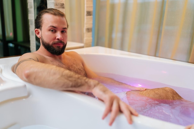 Retrato de un joven apuesto con barba tirado en un jacuzzi disfrutando de un hidromasaje en un salón de spa de lujo mirando a la cámara