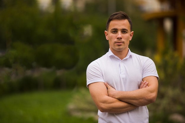 Retrato de un joven apuesto, deportivo y confiado, posando al aire libre con una camiseta blanca de pie con las manos cruzadas. Jardín verde o parque en un fondo borroso. Copiar espacio.