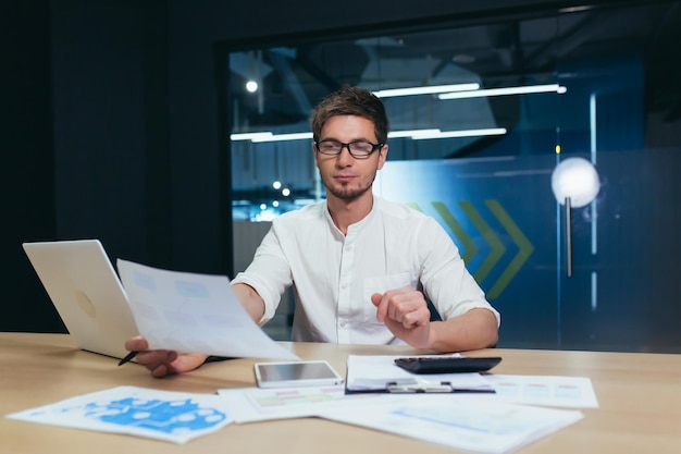 Retrato de un joven apuesto director de contabilidad director de negocios sentado en la oficina en el escritorio con anteojos trabajando en una computadora portátil con una calculadora y documentos