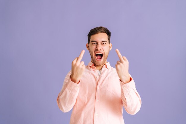 Retrato de un joven apuesto y enojado que muestra alrededor de los dedos medios, demostrando protesta con un gesto grosero y descortés de falta de respeto, rechazando la comunicación sobre un fondo rosa aislado en el estudio.