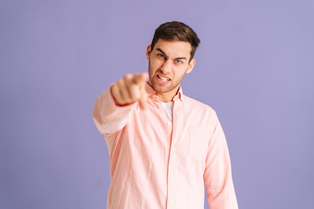 Retrato de un joven apuesto y enojado que te señala con el dedo índice en un fondo morado aislado, mirando la cámara. Un tipo estricto molesto señalando con el dedo índice a la cámara eligiéndote.