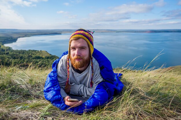 Retrato de un joven y apuesto excursionista de barba pelirroja con un divertido sombrero de yak de lana tejido en Nepal en la naturaleza acostado en un saco de dormir azul paisaje lago y colinas