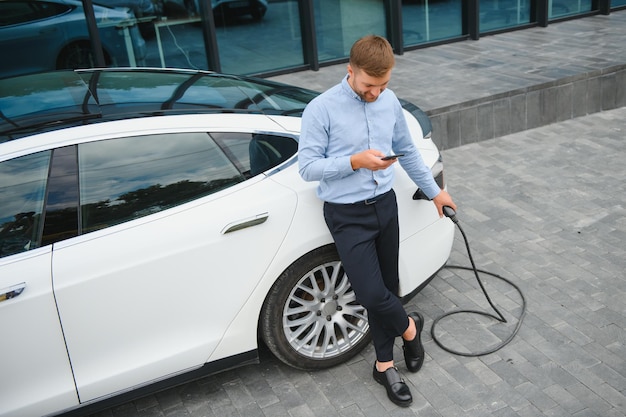 Retrato de un joven apuesto hombre barbudo con ropa informal parado en la estación de carga y sosteniendo un enchufe del cargador para un coche eléctrico Concepto de coche eléctrico ecológico