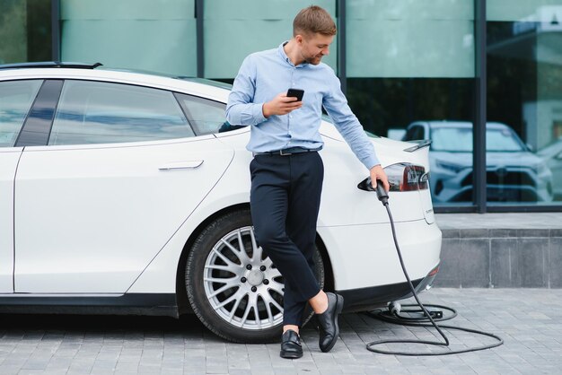 Retrato de un joven apuesto hombre barbudo con ropa informal parado en la estación de carga y sosteniendo un enchufe del cargador para un coche eléctrico Concepto de coche eléctrico ecológico