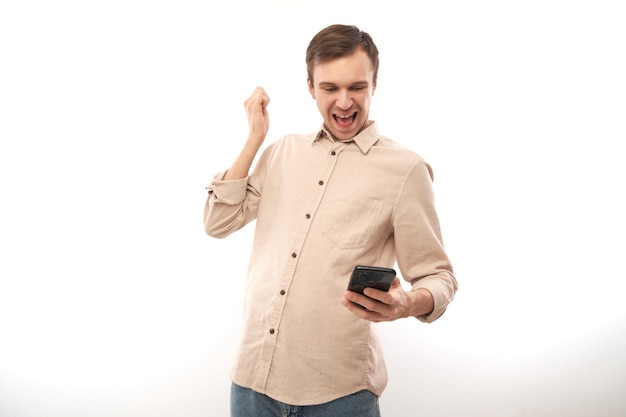 Retrato de joven apuesto hombre caucásico usando teléfono móvil y sonriendo apretando el puño celebrando la victoria aislado en el fondo blanco del estudio expresión facial alegre