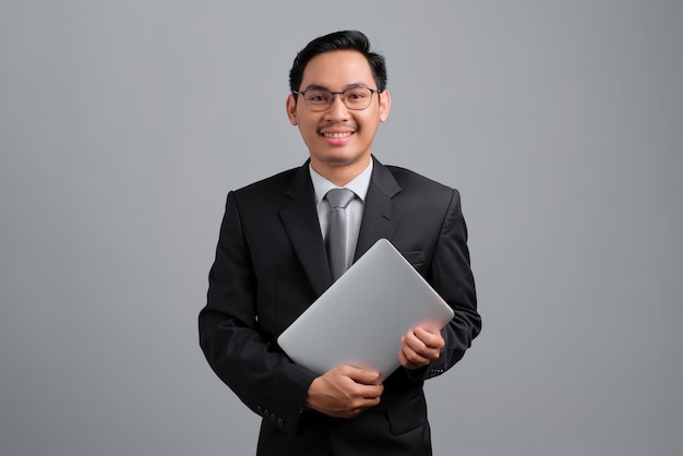 Retrato de un joven y apuesto hombre de negocios sonriente con traje formal y gafas sosteniendo una laptop cerrada aislada de fondo gris