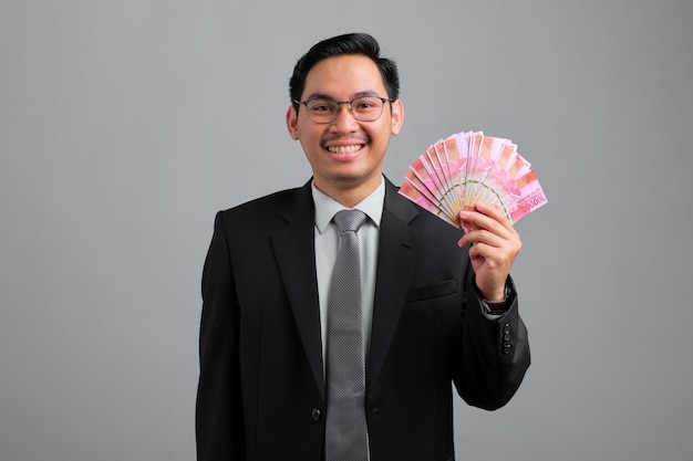 Retrato de un joven y apuesto hombre de negocios sonriente con traje formal que sostiene un abanico de billetes en manos aisladas de fondo gris