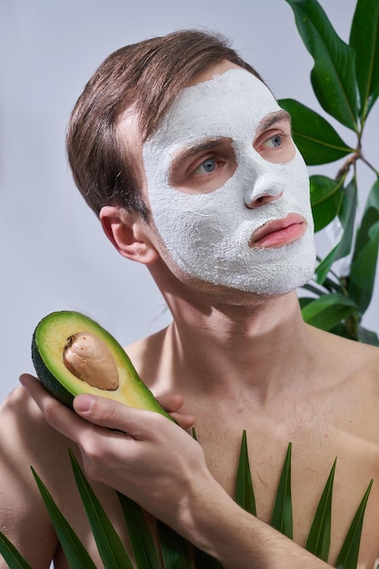 Retrato de un joven apuesto con máscara cosmética en la cara sosteniendo aguacate en la mano aislado en un fondo de hojas de palma exóticas
