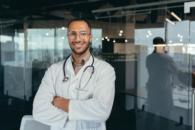 Retrato de un joven y apuesto médico hispano de pie en la sala de cristal del hospital vistiendo