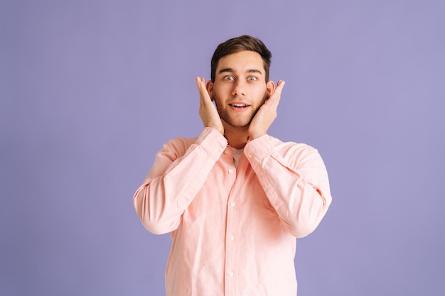 Retrato de un joven apuesto que cubre las orejas con las manos no quiere escuchar de pie sobre un fondo rosa aislado en el estudio mirando la cámara. Concepto de silencio.