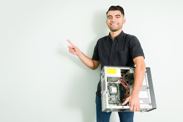 Retrato de un joven apuesto que sonríe mientras lleva una computadora CPU y hardware mientras señala una pared de espacio en blanco en su taller de reparación