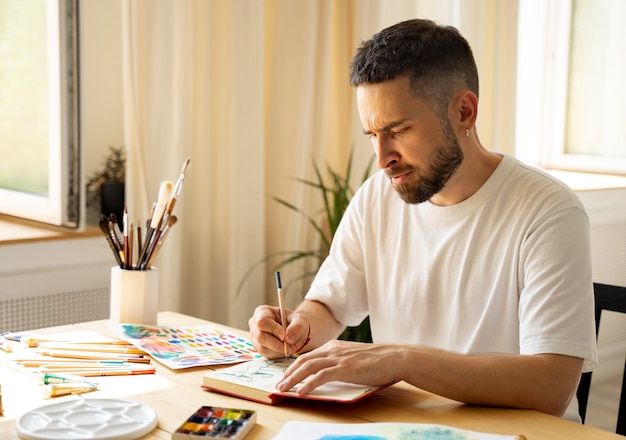 Foto retrato de joven artista masculino blanco caucásico concentrado de pelo oscuro con barba en una camiseta blanca. luz del sol. sentado en un estudio en la mesa de madera. en la mano sosteniendo un lápiz de color. diy.